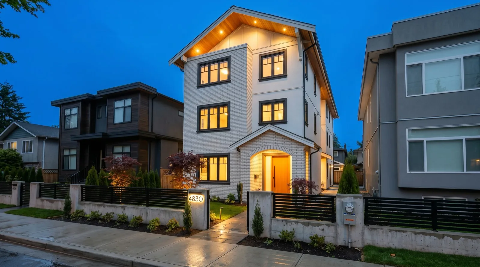 Newly completed three-storey Vancouver multiplex photographed at blue hour on a narrow residential lot, white brick facade with dark window trim, peaked roof with warm illuminated cedar soffit, arched entry with a glowing wood front door, modern horizontal slatted black metal front gate with illuminated address number, low concrete retaining wall with Japanese maples and step-lighting, a BC Hydro service meter visible at the side gangway, neighbouring contemporary duplexes of mixed styles on either side